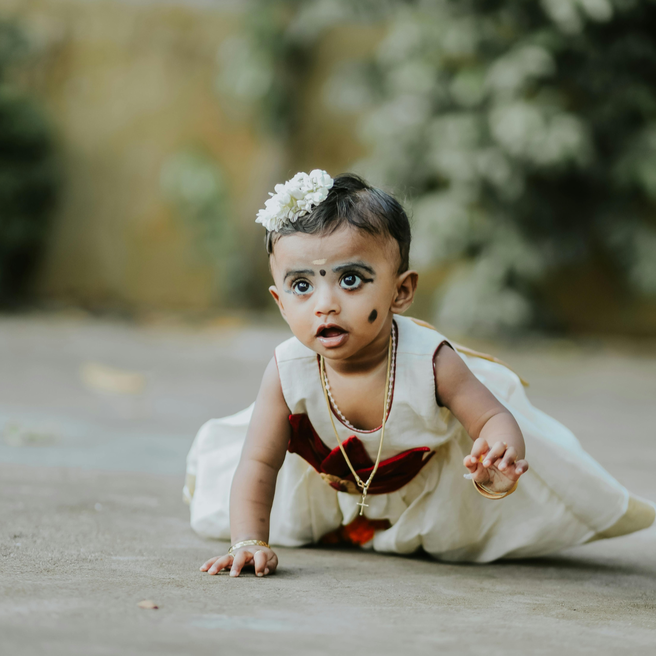 Baby in a white dress with red accents and a flower in hair, sitting on a path with greenery.